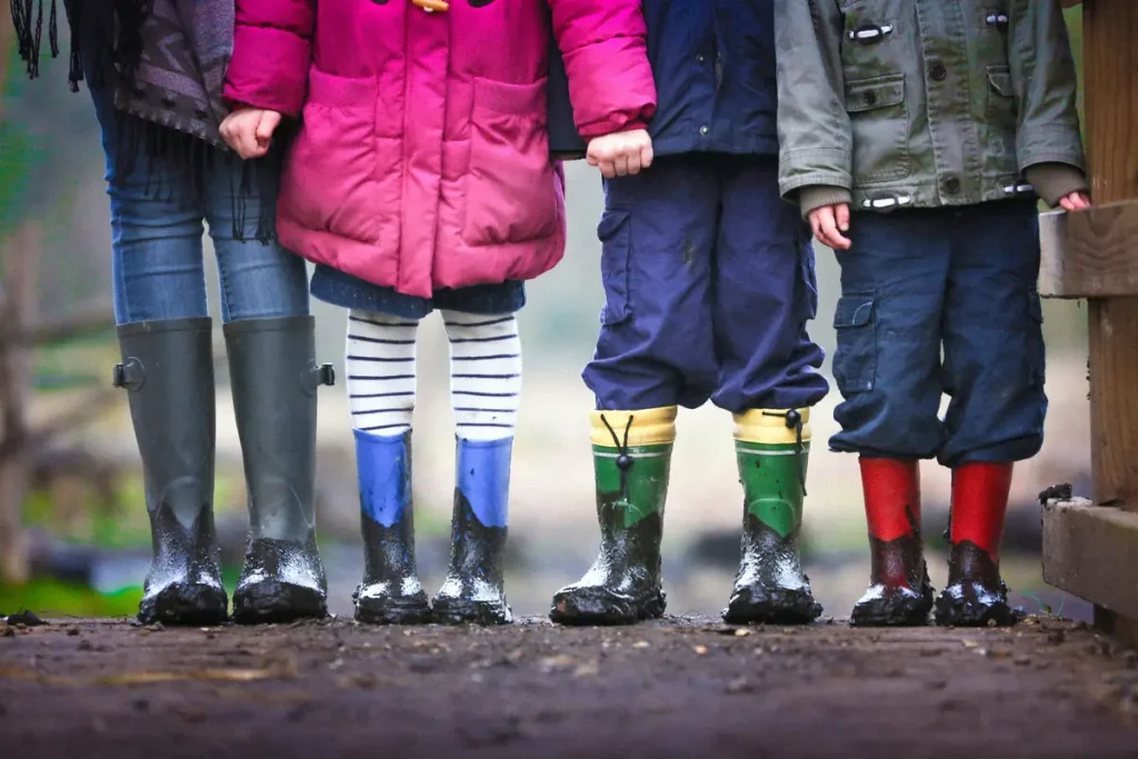 Four children wearing wellies and coats stand side by side on a muddy path, shown from the waist down.