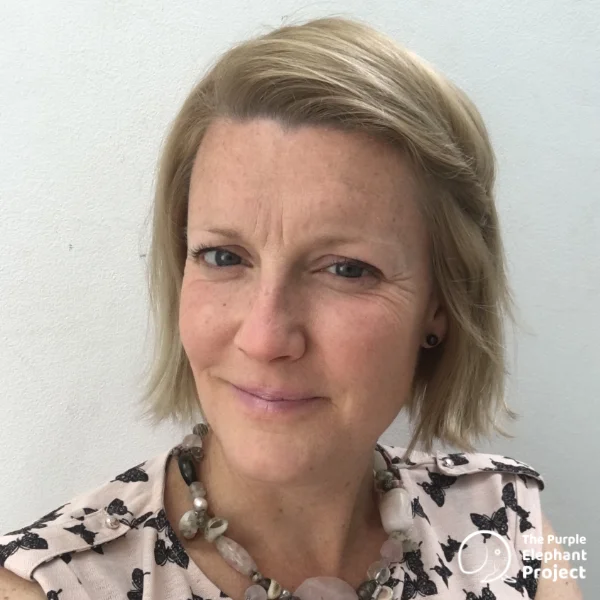 A woman with short blonde hair, wearing a patterned top and a beaded necklace, stands in front of a plain white background. The Purple Elephant Project logo is in the bottom right corner.