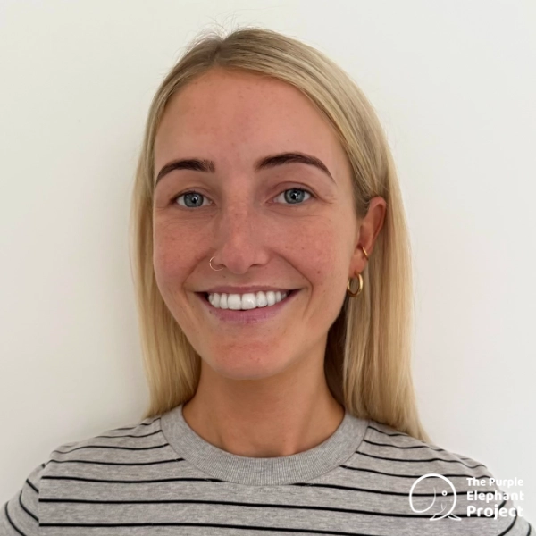 Woman with long blonde hair, wearing a striped shirt, smiling in front of a plain white background. The Purple Elephant Project logo is in the lower right corner.