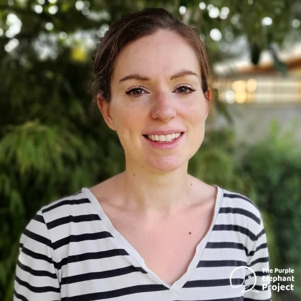 A woman in a black and white striped shirt stands outdoors, smiling at the camera with greenery in the background. The Purple Elephant Project logo is in the bottom right-hand corner.