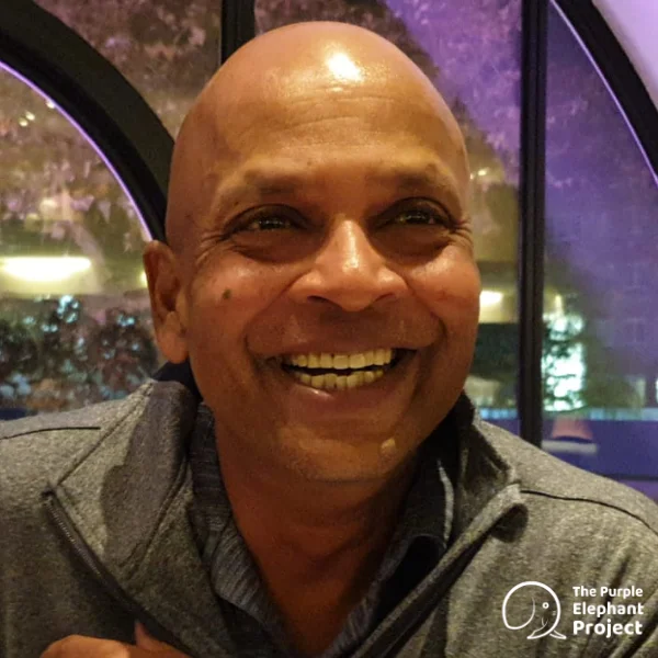 A smiling bald man in a grey jacket sits indoors with purple lighting. The Purple Elephant Project logo is in the bottom right corner.