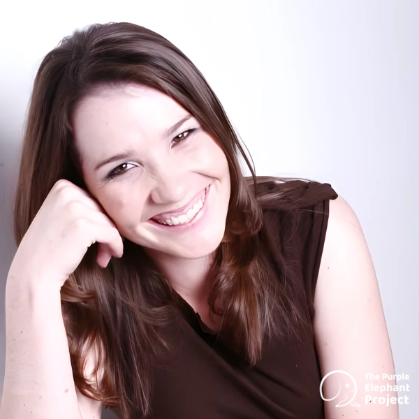 A woman with long brown hair, wearing a dark sleeveless top, smiles whilst resting her head on her hand. The Purple Elephant Project logo is visible in the bottom right corner.