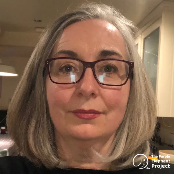 Middle-aged woman with grey hair and glasses looks at the camera, standing indoors. The Purple Elephant Project logo is visible in the lower right corner.
