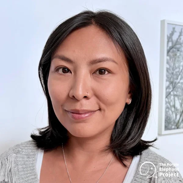 Sibal Cheung, a woman with straight black hair and a light jumper smiles softly at the camera. There is a white wall and a framed picture in the background. The Purple Elephant Project logo is visible.