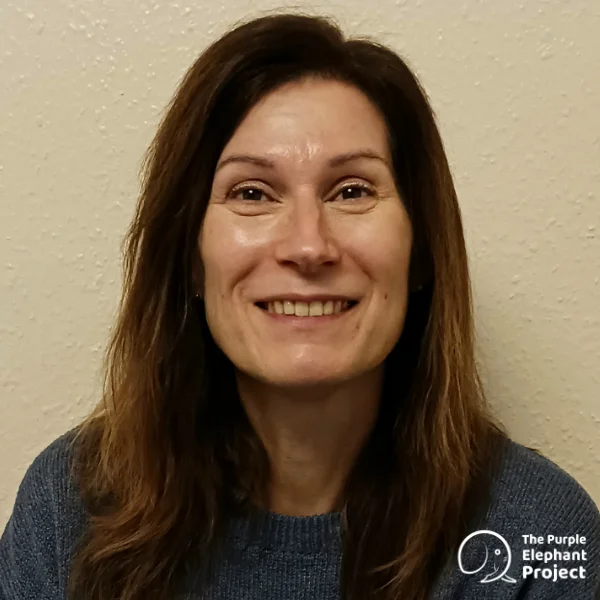Woman with long brown hair smiles at the camera, wearing a blue jumper. A white The Purple Elephant Project logo is visible in the lower right corner.