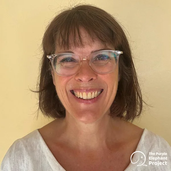 Smiling woman with short brown hair and glasses, wearing a white top, stands against a plain light background. The Purple Elephant Project logo is in the bottom right corner.