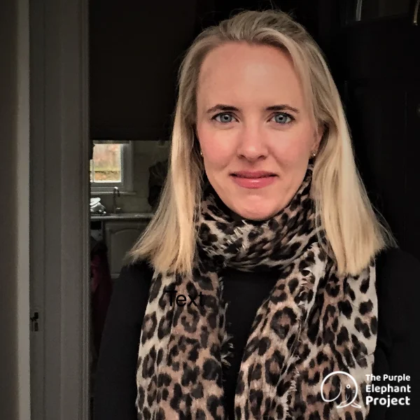 A woman with straight blonde hair wears a leopard print scarf and black top, standing indoors near a door. The Purple Elephant Project logo appears in the lower right corner.