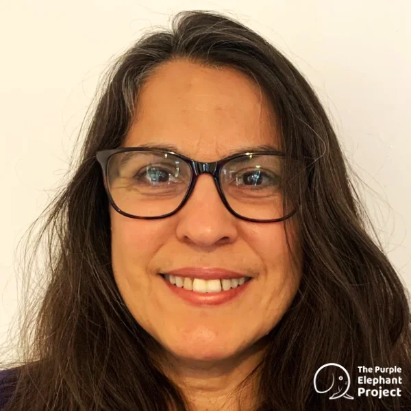 A woman with long brown hair and glasses smiles at the camera. She is standing in front of a plain white background. The Purple Elephant Project logo is in the bottom right corner.