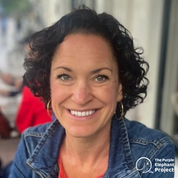 Woman with short curly hair smiles at the camera, wearing a denim jacket. The Purple Elephant Project logo is in the bottom right corner.