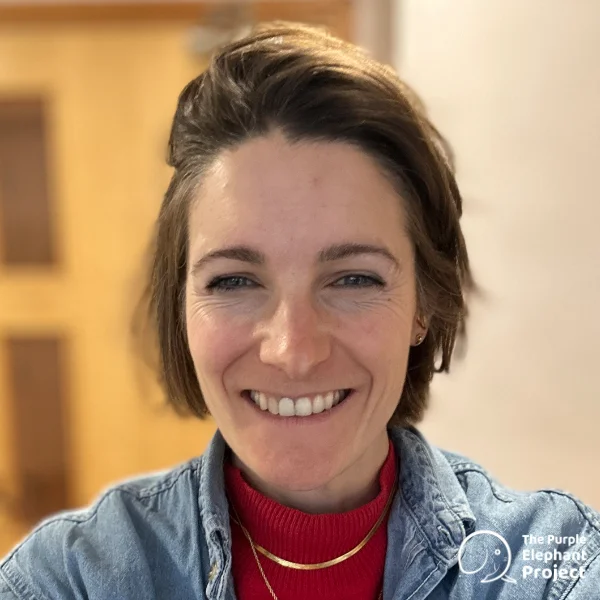 A woman with short brown hair smiles at the camera, wearing a red polo neck and a denim jacket. A logo in the corner reads The Purple Elephant Project.