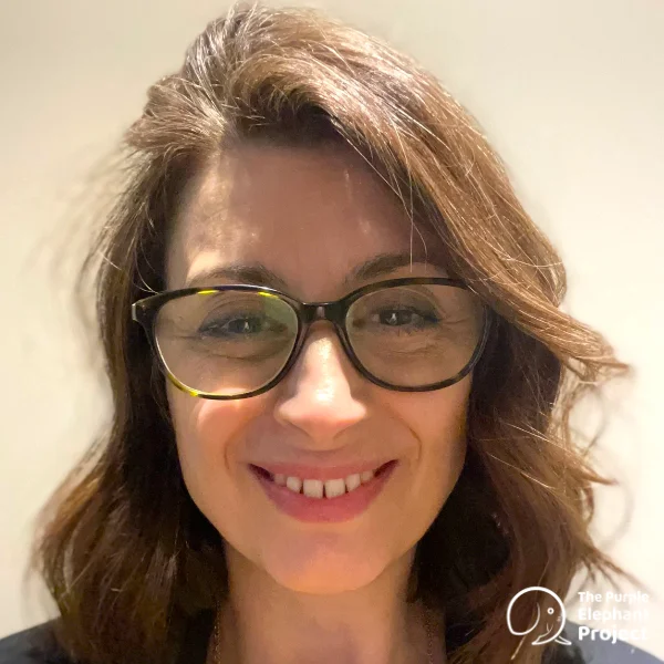 A woman with shoulder-length brown hair and glasses smiles at the camera in front of a plain background.