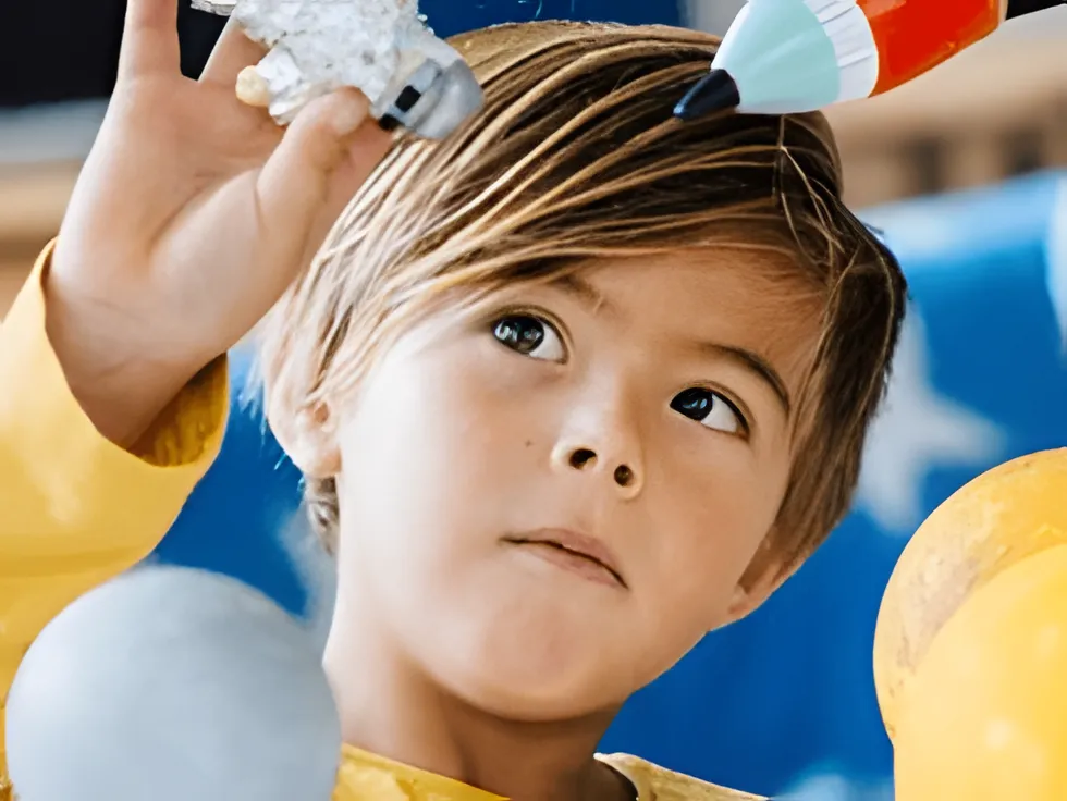 A young child in a yellow shirt holds up a toy astronaut and a toy rocket, looking at them with concentration. Blue and yellow decorations are visible in the background.