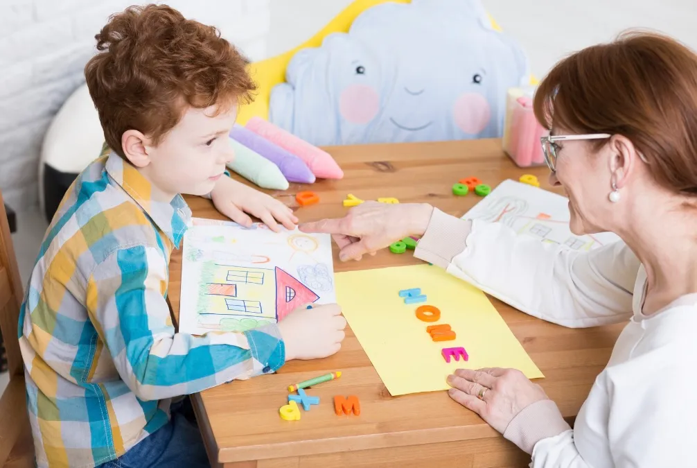 A young boy shows his drawing to an adult woman at a table. The woman points to colourful letters spelling HOME on a yellow paper beside art supplies and magnetic letters.