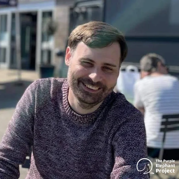 A man with brown hair and a beard sits outside at a table, smiling. He wears a maroon jumper. The logo “The Purple Elephant Project” is in the bottom right corner.