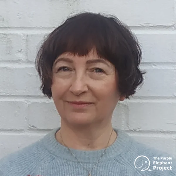 A person with short brown hair and a grey jumper stands in front of a white brick wall. The Purple Elephant Project logo is in the bottom right corner.