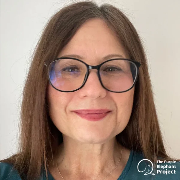 A woman with straight brown hair and glasses smiles at the camera. She is wearing a green shirt. The Purple Elephant Project logo appears in the bottom right corner.