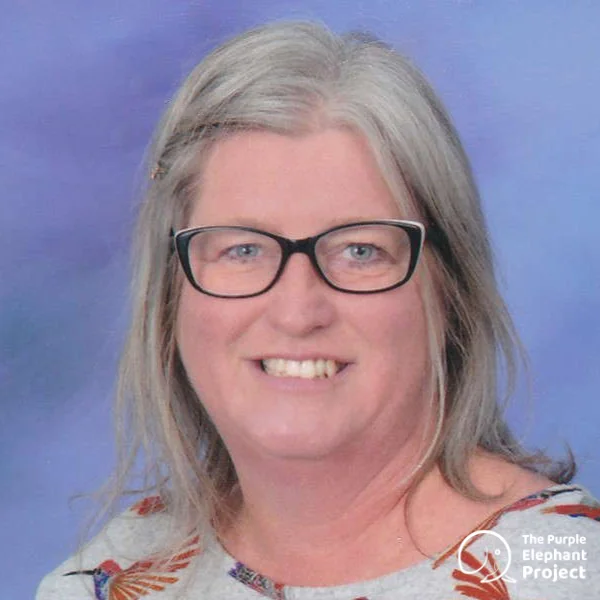 A woman with grey hair and glasses smiles at the camera, wearing a patterned top. A logo in the corner reads The Purple Elephant Project.