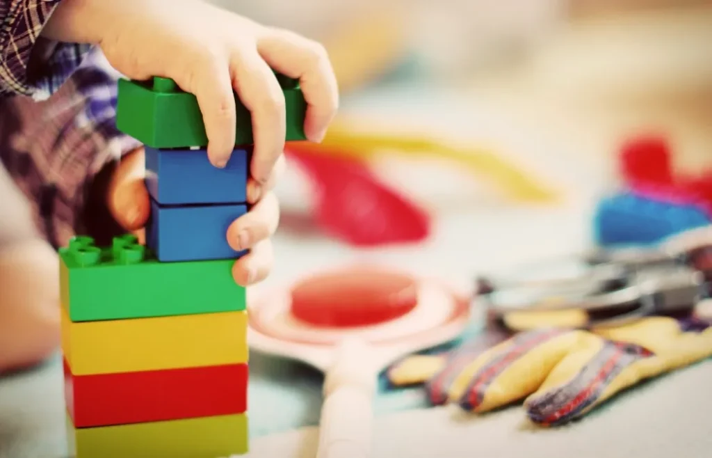 A child’s hand stacks colourful toy blocks on a surface, with other toys blurred in the background.