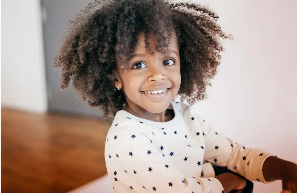 Smiling young child with curly hair wears a white shirt with black stars, sitting indoors with a blurred brown and white background.