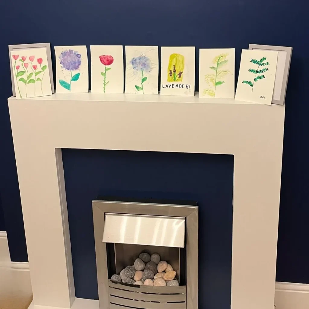 A white mantelpiece over a modern fireplace displays seven small, hand-painted flower artworks against a dark blue wall.