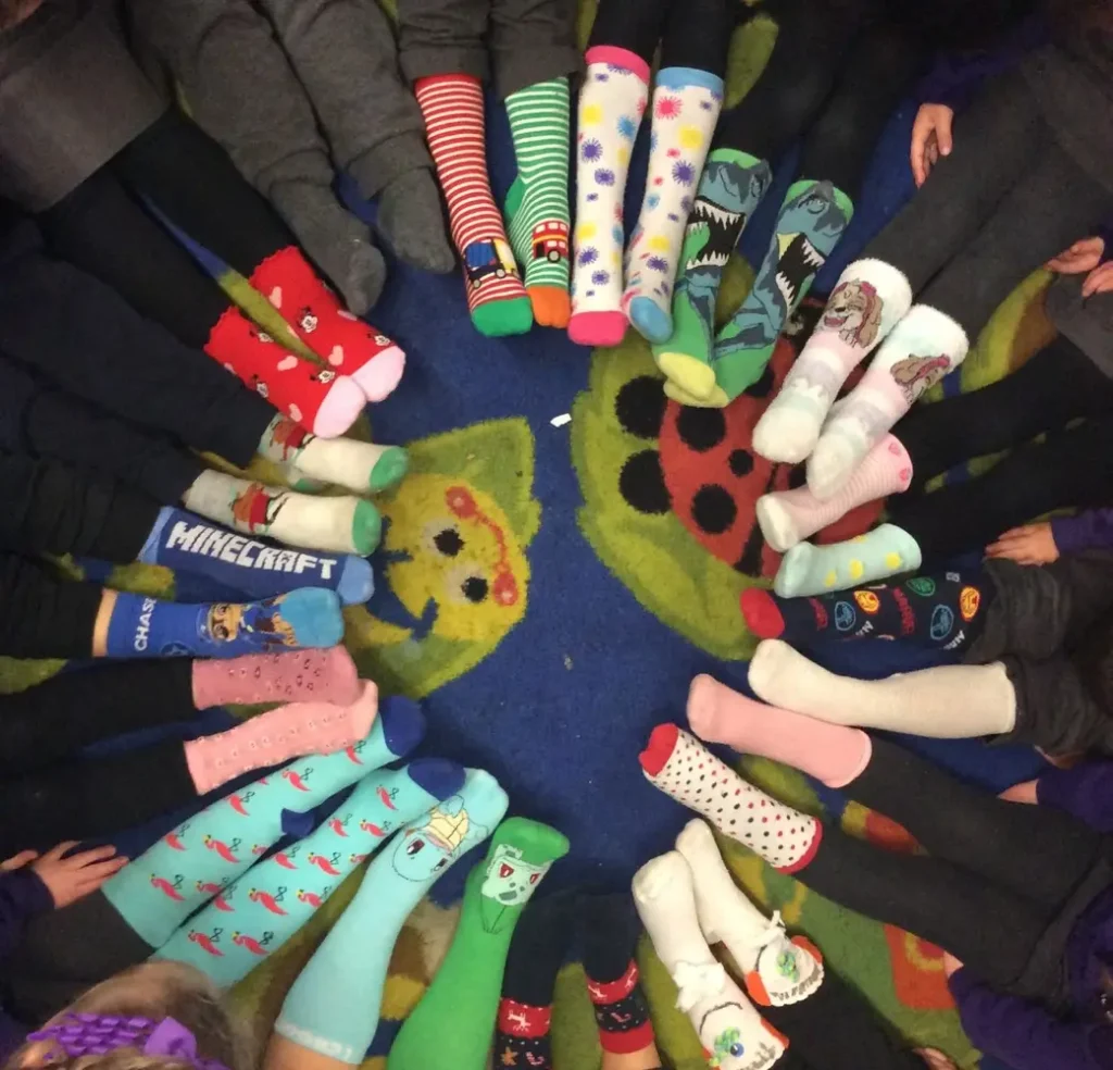 Children sit in a circle, feet extended to the centre, wearing various colourful and patterned socks, on a carpet with ladybird and flower designs.