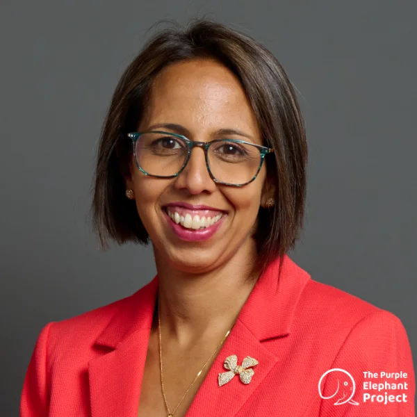 Woman with straight brown hair, glasses, and coral blazer smiling at the camera in front of a plain grey background; The Purple Elephant Project logo is in the image corner.