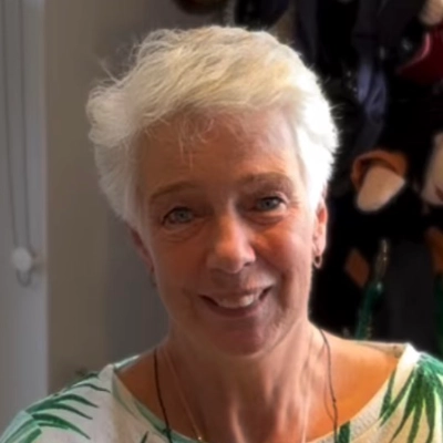 An older woman with short white hair smiles at the camera indoors. She wears a light printed top and a necklace.