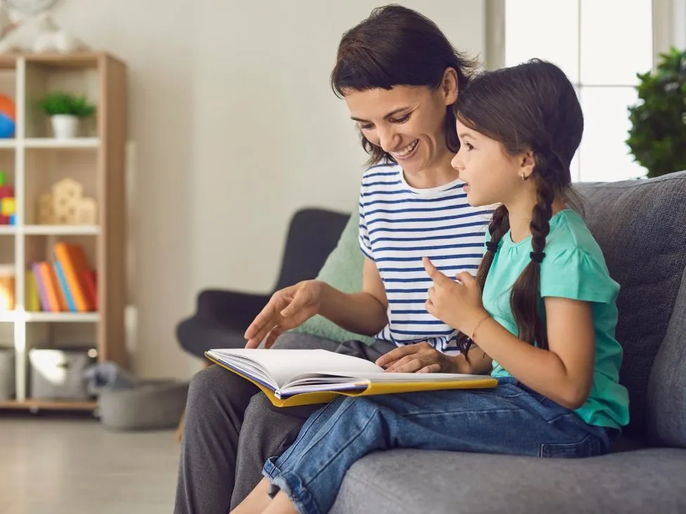 An adult and a child sit on a sofa reading a book together in a brightly lit living room with shelves in the background.