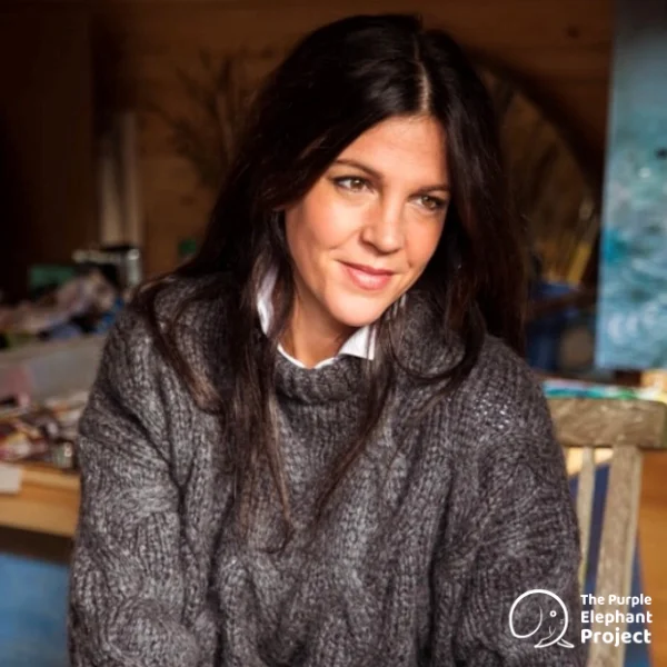 A woman with long dark hair wears a chunky grey jumper and sits indoors, smiling slightly. The Purple Elephant Project logo is visible in the lower right corner.