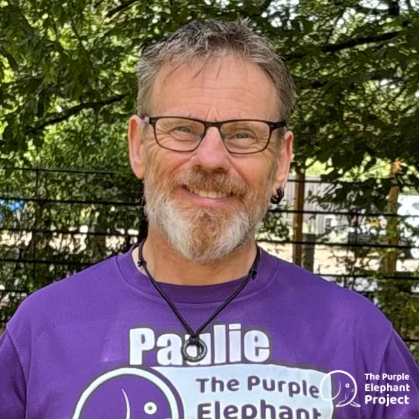 A smiling man with glasses and a beard stands outdoors wearing a purple The Purple Elephant Project shirt. Trees and a metal fence are visible in the background.