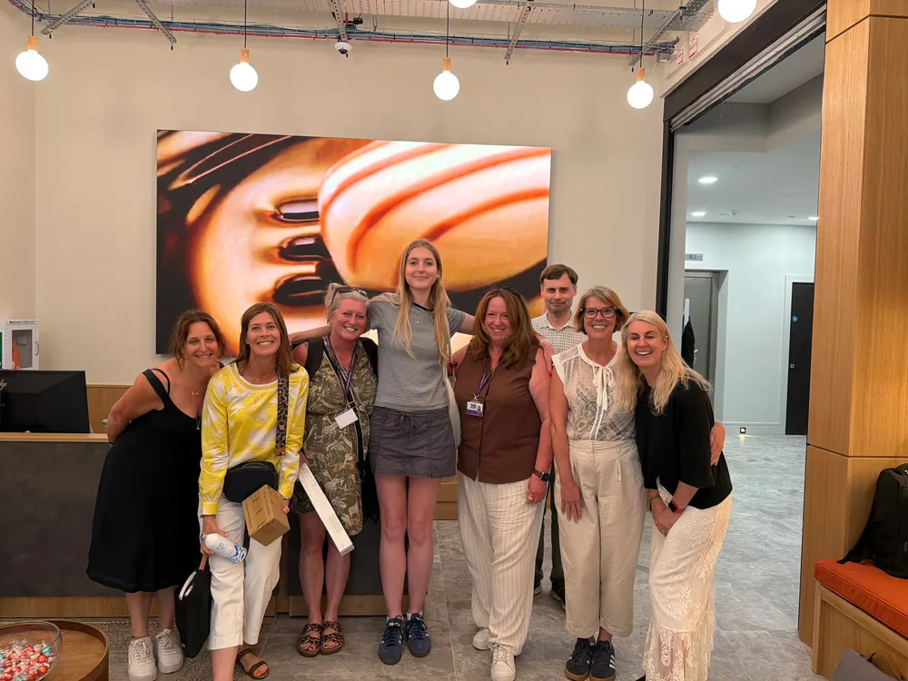 Nine people pose together and smile in an office lobby with modern decor and a large abstract painting on the wall behind them.