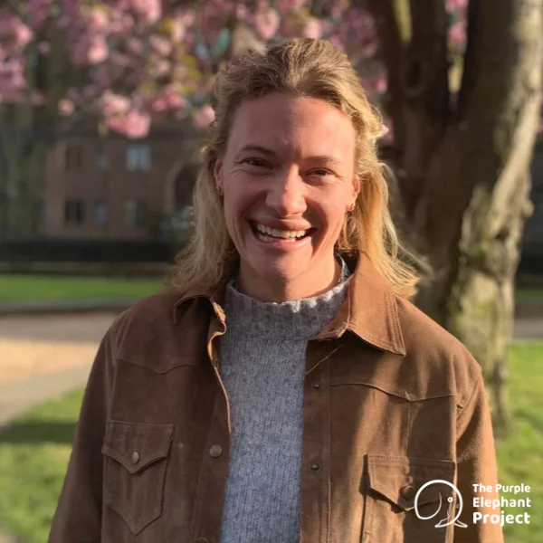 A woman smiling outdoors, wearing a brown jacket over a grey jumper, with trees and a building in the background. The Purple Elephant Project logo is in the bottom right corner.