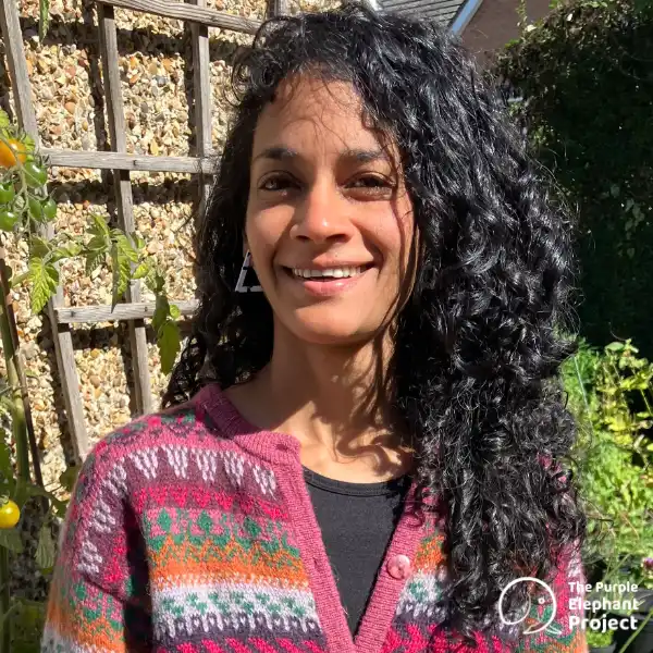 Charlene Fernandes, standing with curly black hair and a patterned jumper smiles outdoors in sunlight, standing near a wooden trellis and stone wall. The Purple Elephant Project logo is visible in the corner.