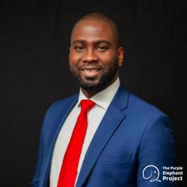 A man in a blue suit, white shirt, and red tie smiles at the camera against a plain dark background. The Purple Elephant Project logo is visible in the corner.