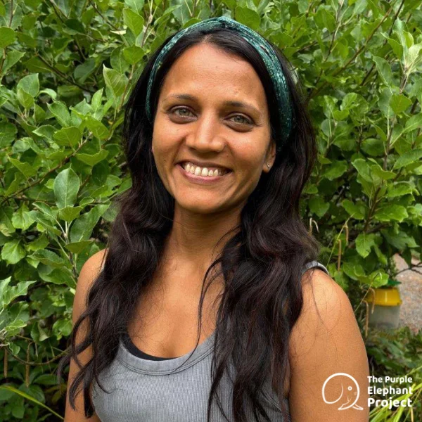 Archana Chatterjee, a woman with long dark hair and a green headband smiles in front of leafy green bushes, wearing a grey vest top. The Purple Elephant Project logo appears in the lower right corner.