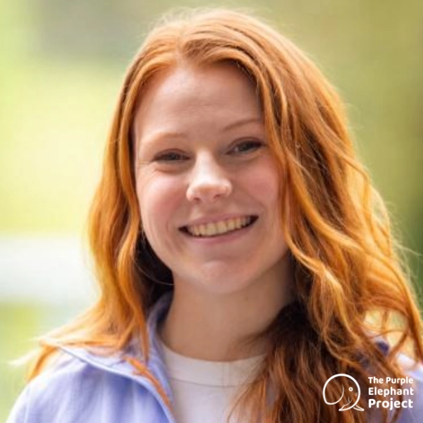 Emily Sheridan, a young woman with long red hair smiles at the camera. She is wearing a light purple jacket and stands in front of a blurred green background. The Purple Elephant Project logo is in the corner.