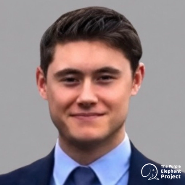 Nicholas Ireland, in a suit and tie, smiling slightly, stands against a plain grey background. The Purple Elephant Project logo is visible in the bottom right-hand corner.