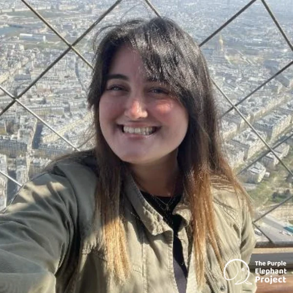 Nicola Mackenzie, smiling in front of a metal fence with a cityscape in the background. The Purple Elephant Project logo appears in the corner.