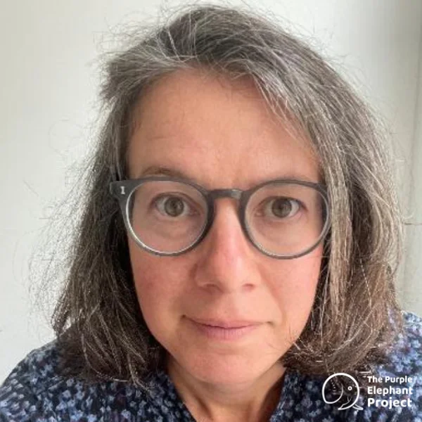 Rosie Creer, a woman with shoulder-length grey hair and round glasses looks at the camera. She is wearing a patterned top. The Purple Elephant Project logo is in the bottom right corner.