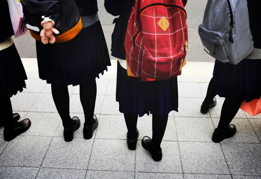 Four students in dark uniforms and rucksacks stand on a tiled pavement; only their lower halves are visible, facing away from the camera.