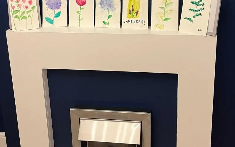 A white mantelpiece over a modern fireplace displays seven small, hand-painted flower artworks against a dark blue wall.