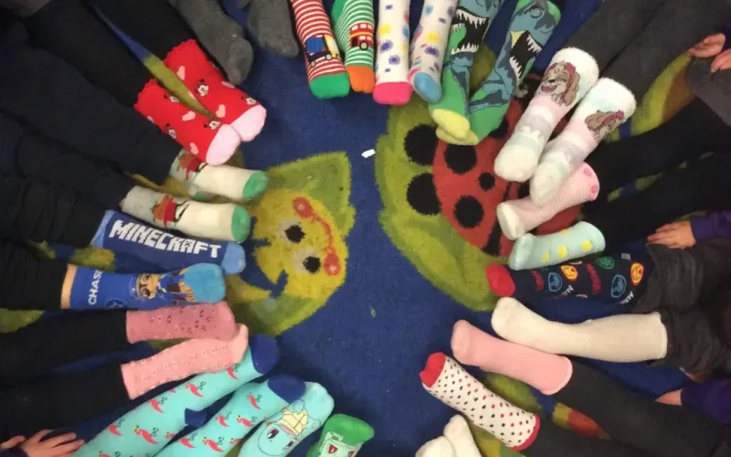 Children sit in a circle, feet extended to the centre, wearing various colourful and patterned socks, on a carpet with ladybird and flower designs.