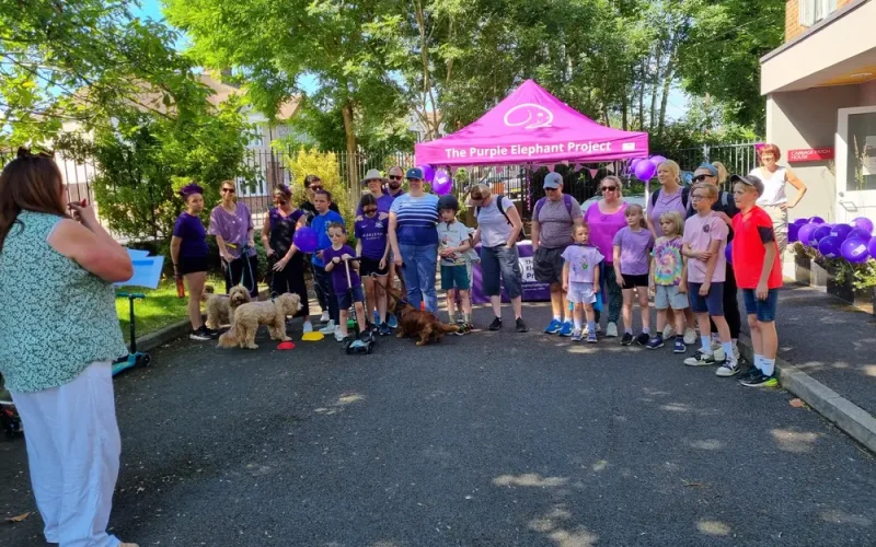 A group of people and dogs pose for a photo near a purple tent labelled The Purple Elephant Project on a sunny day, with someone taking their picture.