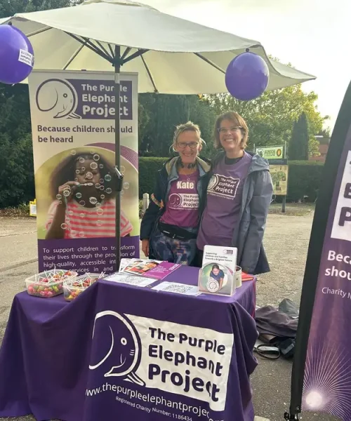 Two people stand behind a table with The Purple Elephant Project banner and materials at an outdoor event, promoting support for children.