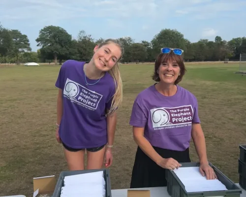 Two women in purple “Elephant Project” shirts stand behind tables with bins of papers, outdoors on grass under a partly cloudy sky. Yellow signs read “NUMBER COLLECTION HERE”.