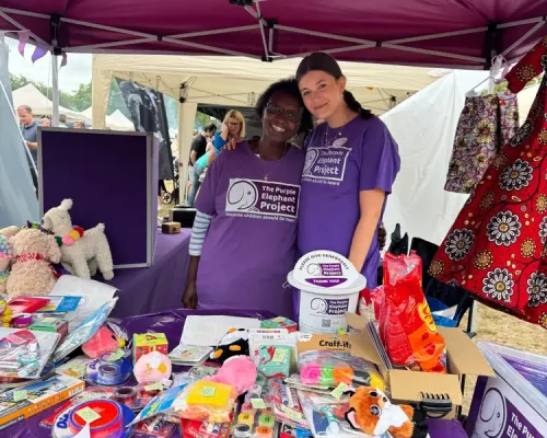 Two women in purple shirts stand behind a fundraising stall with toys and donation boxes under a canopy at an outdoor event.