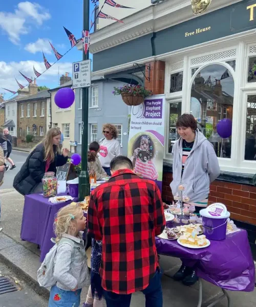 People gather outside a pub at tables with purple decorations and cakes for a charity event. Banners and balloons are visible, and children and adults interact on a cloudy day.