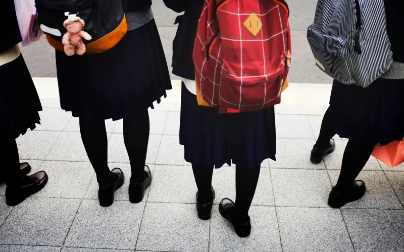Four students in dark uniforms and rucksacks stand on a tiled pavement; only their lower halves are visible, facing away from the camera.