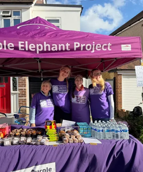 Four people in purple shirts stand behind a table of snacks and drinks under a Purple Elephant Project marquee at an outdoor event in a residential area.